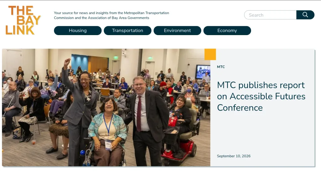A diverse group of people, including individuals using wheelchairs and mobility scooters, pose and smile in a conference room; a presentation screen is visible behind them.
