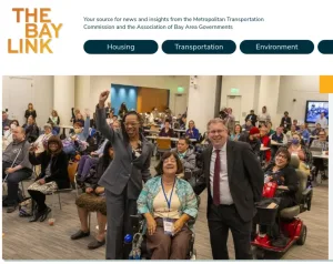 A diverse group of people, including individuals in wheelchairs, smile and cheer at an indoor event. Two people stand at the front, one raising a fist in celebration, under a sign reading THE BAY LINK..